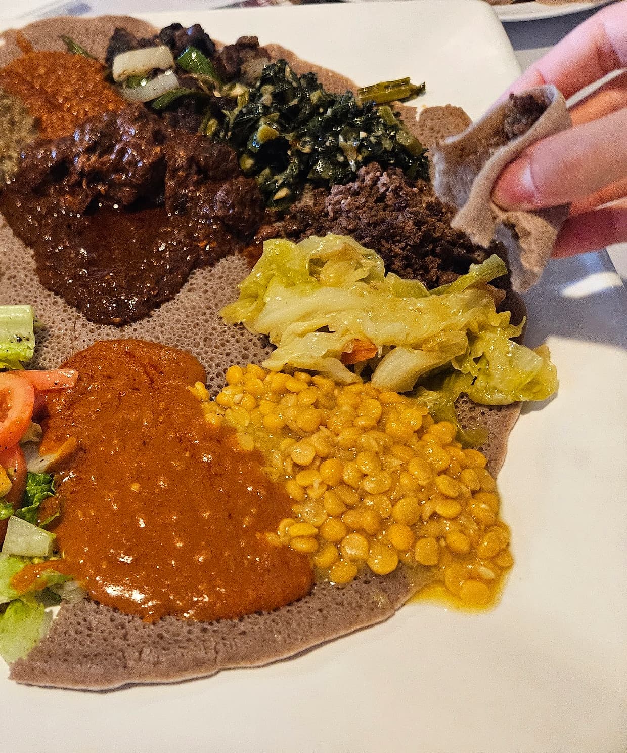 A shared round of injera at Zagol, spread with stews and salad, with a hand tearing off a piece to scoop with